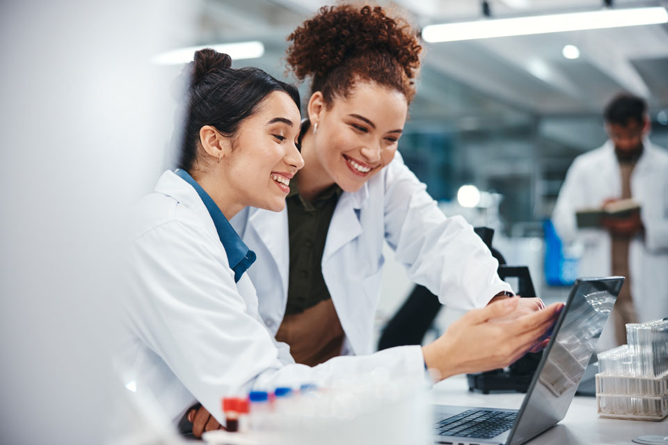 Two people in lab coats smiling and looking at a laptop screen